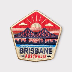 May include: Embroidered patch featuring a bridge over water with a sunset and the text "BRISBANE AUSTRALIA." The patch is in a pentagon shape with a red sky, blue bridge, and a beige border.