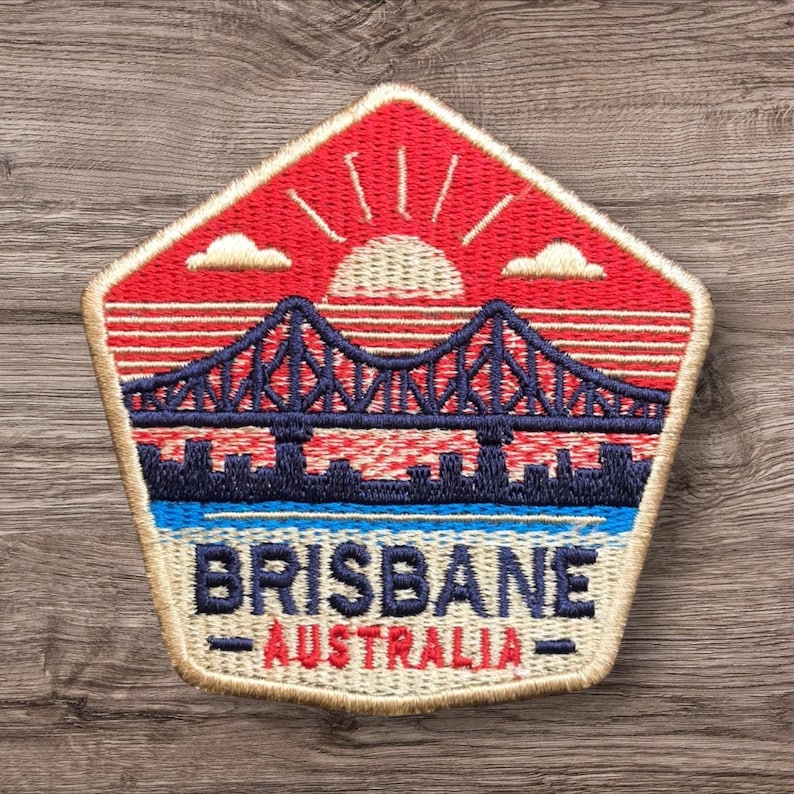 May include: Embroidered patch featuring a bridge over water with a cityscape silhouette. The patch has a red and blue color scheme with the words "BRISBANE AUSTRALIA" in a blue and white font. The patch is in a pentagon shape.