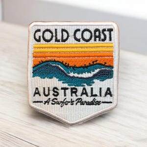 May include: Embroidered patch with the text "Gold Coast Australia" above a sunset design in orange and yellow, and a turquoise ocean wave. The text "A Surfer's Paradise" is below the word Australia.