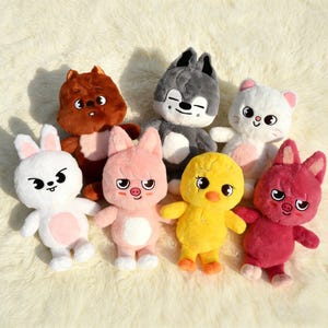 May include: A collection of seven plush animal toys in various colors. The plushies include a brown squirrel, a gray wolf, a white cat, a white rabbit, a pink pig, a yellow duck, and a red pig. Each toy has a unique facial expression.