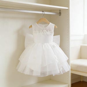 May include: A white sleeveless dress with a layered tulle skirt and a large bow in the back. The bodice is adorned with floral embellishments. The dress hangs on a wooden hanger inside a closet.