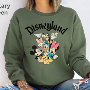 May include: Military green sweatshirt featuring the word "Disneyland" in a black script font, with a graphic of Disney characters. The sweatshirt also has the text "EST. 1955".