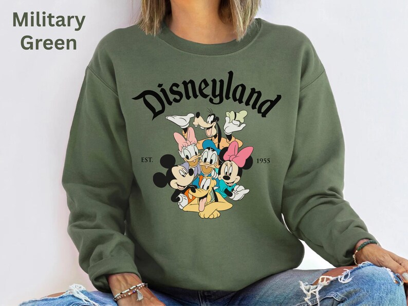 May include: Military green sweatshirt featuring the word "Disneyland" in a black script font, with a graphic of Disney characters. The sweatshirt also has the text "EST. 1955".