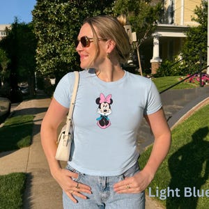 May include: Light blue t-shirt with a Minnie Mouse graphic. The cartoon character is shown with a pink bow, dress, and shoes, and a blue scarf. The shirt is worn with blue jeans.