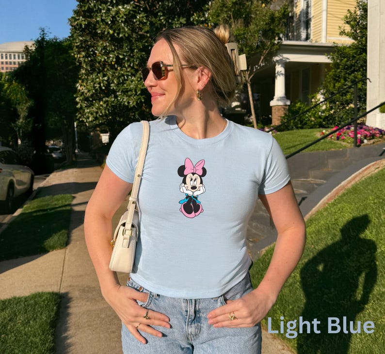 May include: Light blue t-shirt with a Minnie Mouse graphic. The cartoon character is shown with a pink bow, dress, and shoes, and a blue scarf. The shirt is worn with blue jeans.