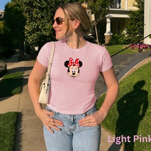 May include: Light pink t-shirt featuring a Minnie Mouse graphic with a red bow and black ears. The shirt is a crew neck and short-sleeved. The text "Light Pink" is at the bottom right of the image.