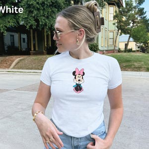 May include: White t-shirt with a Minnie Mouse graphic. The cartoon character has a pink bow, black ears, and a blue scarf. The word "White" is printed in the upper left corner of the image.