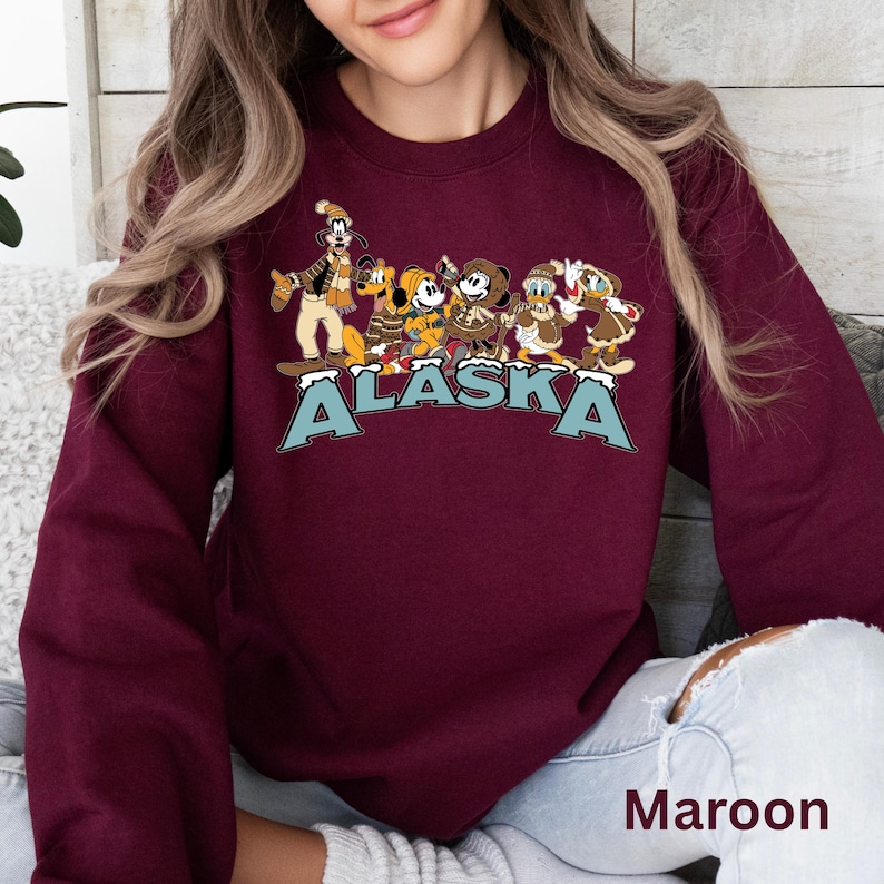 Peut inclure: Sweat-shirt bordeaux avec un motif de personnages de dessins anim&eacute;s en v&ecirc;tements d'hiver et le mot "ALASKA" en bleu sarcelle. Le sweat-shirt est de couleur bordeaux unie.