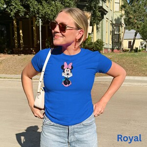 May include: Royal blue t-shirt with a Minnie Mouse graphic, including a pink bow and skirt. The shirt has short sleeves and a crew neck. The word "Royal" is printed in blue at the bottom of the image. The shirt is worn with light-wash denim shorts.
