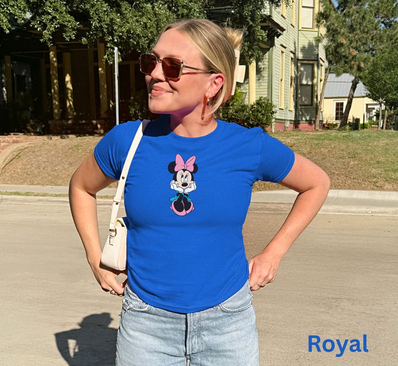 May include: Royal blue t-shirt with a Minnie Mouse graphic, including a pink bow and skirt. The shirt has short sleeves and a crew neck. The word "Royal" is printed in blue at the bottom of the image. The shirt is worn with light-wash denim shorts.