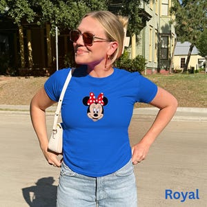 May include: Royal blue t-shirt featuring a Minnie Mouse graphic with a red and white polka dot bow. The shirt is a classic crew neck style and is paired with light wash denim jeans. The word "Royal" is printed in blue on the bottom right.