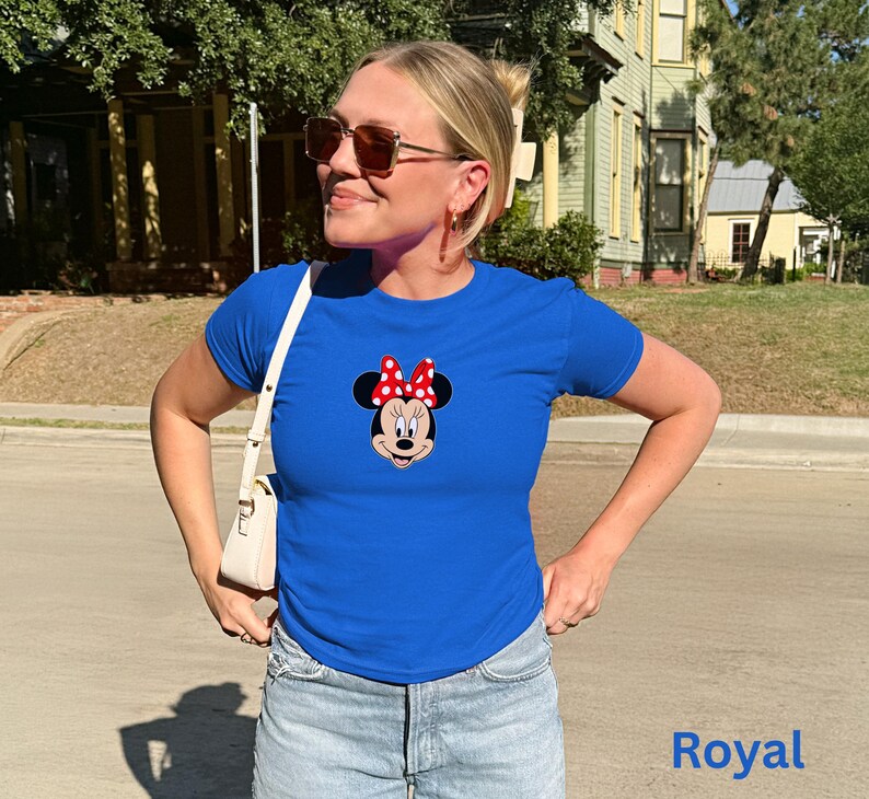 May include: Royal blue t-shirt featuring a Minnie Mouse graphic with a red and white polka dot bow. The shirt is a classic crew neck style and is paired with light wash denim jeans. The word "Royal" is printed in blue on the bottom right.