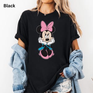 May include: Black t-shirt featuring a Minnie Mouse graphic. The cartoon character is depicted with a pink bow, blue dress, and pink shoes. The word "Black" is printed in the upper left corner of the image.