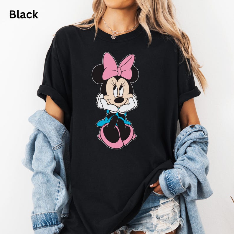 May include: Black t-shirt featuring a Minnie Mouse graphic. The cartoon character is depicted with a pink bow, blue dress, and pink shoes. The word "Black" is printed in the upper left corner of the image.