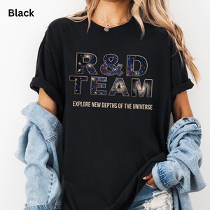 May include: Black t-shirt with the text "R&D TEAM" in a gold outline, with a galaxy design filling the letters. The text "EXPLORE NEW DEPTHS OF THE UNIVERSE" is below the main text. The shirt is worn with a denim jacket.
