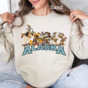 Peut inclure: Sweat-shirt couleur sable avec un motif de personnages de dessins anim&eacute;s en v&ecirc;tements d'hiver et le mot "ALASKA" en lettres bleues. Les personnages incluent Dingo, Pluto, Mickey Mouse, Minnie Mouse, Donald Duck et Daisy Duck.