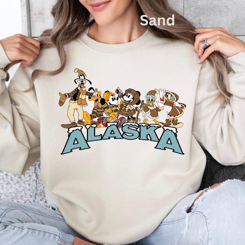 Peut inclure: Sweat-shirt couleur sable avec un motif de personnages de dessins anim&eacute;s en v&ecirc;tements d'hiver et le mot "ALASKA" en lettres bleues. Les personnages incluent Dingo, Pluto, Mickey Mouse, Minnie Mouse, Donald Duck et Daisy Duck.