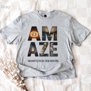 May include: Sport gray t-shirt with the word "AMAZE" in a bold, space-themed design. The shirt features the text "UNCHARTED PATHS. NEW HORIZONS." in a smaller font. The shirt is laid flat on a white surface.