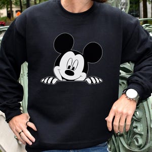 May include: Black crewneck sweatshirt featuring a large, smiling Mickey Mouse graphic in black and white. The iconic cartoon character is peeking over a white edge. The sweatshirt has long sleeves and a classic fit.