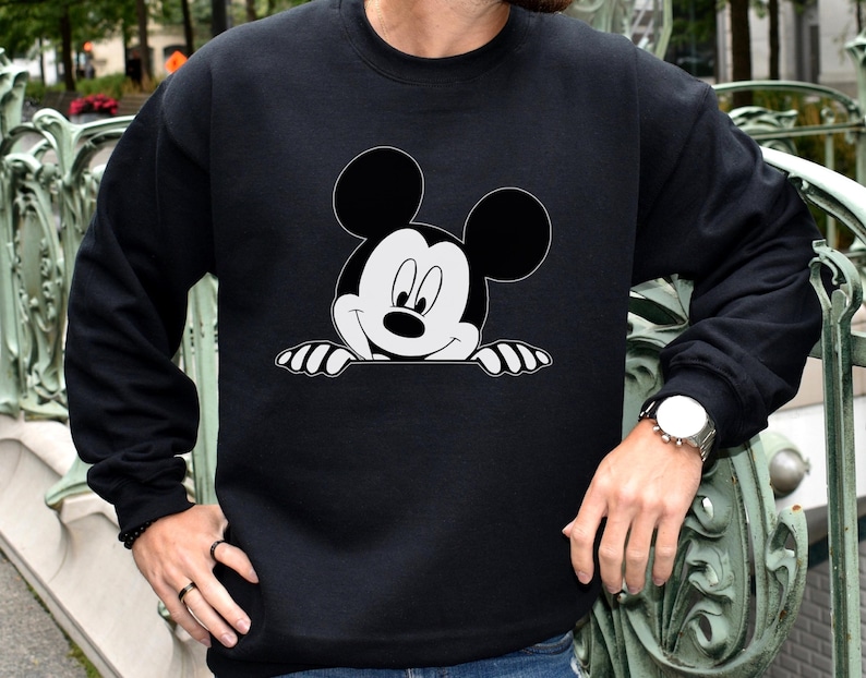 May include: Black crewneck sweatshirt featuring a large, smiling Mickey Mouse graphic in black and white. The iconic cartoon character is peeking over a white edge. The sweatshirt has long sleeves and a classic fit.
