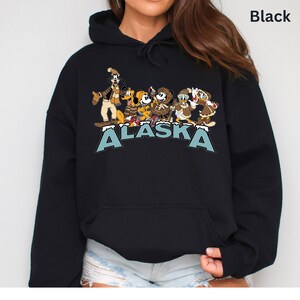 Peut inclure: Sweat-shirt &agrave; capuche noir avec un motif de personnages de dessins anim&eacute;s en v&ecirc;tements d'hiver au-dessus du mot "ALASKA" en lettres turquoise. Le sweat-shirt a une poche avant et une capuche &agrave; cordon.