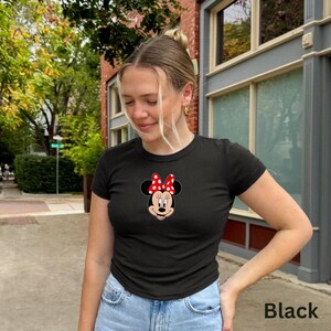 May include: Black short-sleeved t-shirt featuring a Minnie Mouse graphic. The graphic shows Minnie's face with a red and white polka dot bow. The shirt is paired with blue jeans. The word "Black" is printed in the bottom right corner.
