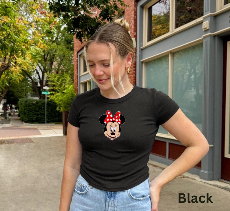 May include: Black short-sleeved t-shirt featuring a Minnie Mouse graphic. The graphic shows Minnie's face with a red and white polka dot bow. The shirt is paired with blue jeans. The word "Black" is printed in the bottom right corner.
