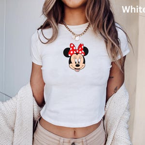 May include: White crop top featuring a Minnie Mouse graphic. The graphic shows Minnie's face with a red bow with white polka dots. The shirt has short sleeves and a round neckline. The word "White" is printed in the upper right corner.