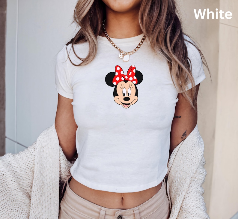 May include: White crop top featuring a Minnie Mouse graphic. The graphic shows Minnie's face with a red bow with white polka dots. The shirt has short sleeves and a round neckline. The word "White" is printed in the upper right corner.