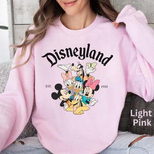 May include: Light pink Disneyland sweatshirt featuring Mickey Mouse, Minnie Mouse, Donald Duck, Daisy Duck, Goofy, and Pluto. The word "Disneyland" is in a black script font above the characters, with "EST. 1955" below.