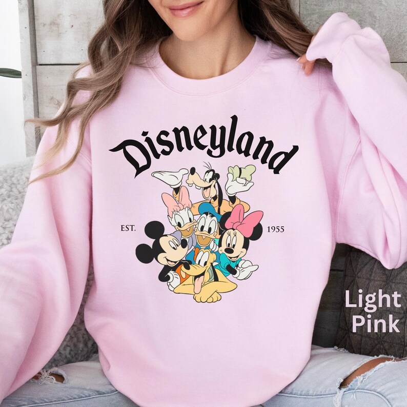 May include: Light pink Disneyland sweatshirt featuring Mickey Mouse, Minnie Mouse, Donald Duck, Daisy Duck, Goofy, and Pluto. The word "Disneyland" is in a black script font above the characters, with "EST. 1955" below.