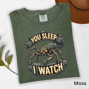 May include: A moss green t-shirt featuring a spider-like creature design on a retro phone. The text "YOU SLEEP" and "I WATCH" are displayed. The shirt is made of a soft material. The t-shirt is a comfortable fit.