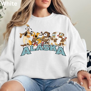 Peut inclure: Sweat-shirt blanc avec un motif de personnages de dessins anim&eacute;s en v&ecirc;tements d'hiver et le mot "ALASKA" en lettres bleues. Les personnages incluent Dingo, Pluto, Mickey Mouse, Minnie Mouse, Donald Duck et Daisy Duck.