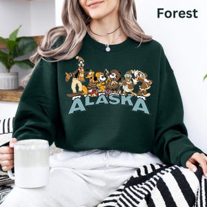 Peut inclure: Un sweat-shirt vert for&ecirc;t avec un motif de personnages de dessins anim&eacute;s et le mot "ALASKA" en bleu et blanc. Le sweat-shirt a un col rond et des manches longues. Le mot "Forest" est imprim&eacute; en blanc en haut &agrave; droite.