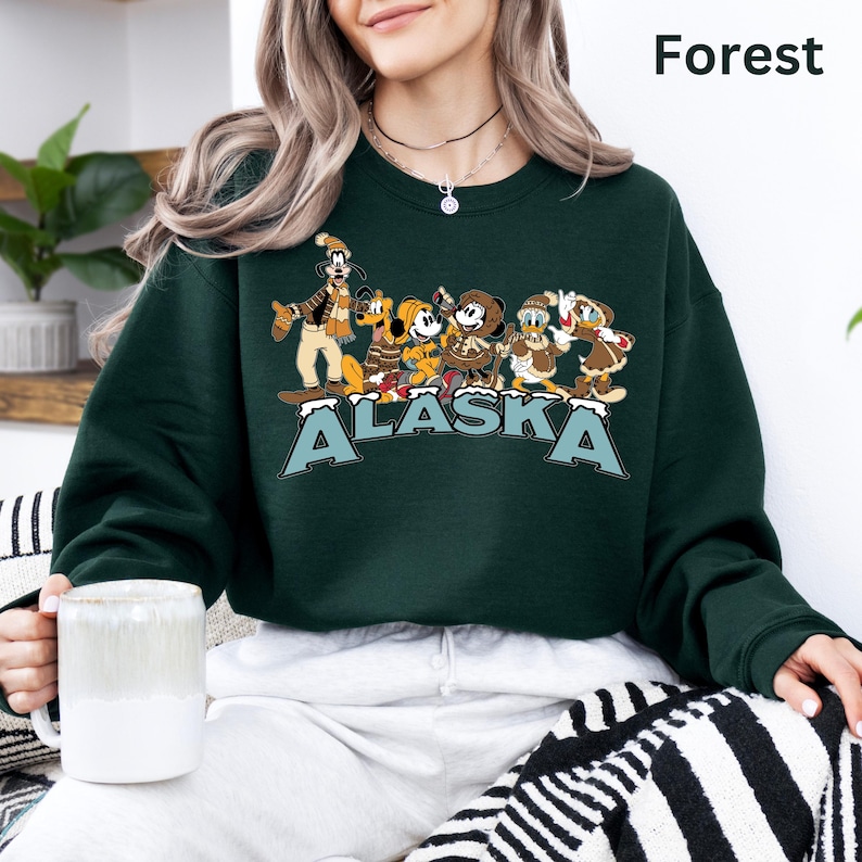 Peut inclure: Un sweat-shirt vert for&ecirc;t avec un motif de personnages de dessins anim&eacute;s et le mot "ALASKA" en bleu et blanc. Le sweat-shirt a un col rond et des manches longues. Le mot "Forest" est imprim&eacute; en blanc en haut &agrave; droite.