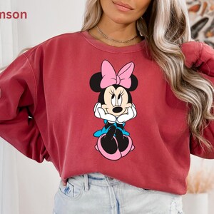 May include: Crimson-colored sweatshirt featuring a Minnie Mouse graphic. The graphic shows Minnie Mouse with a pink bow, blue accents, and black ears. The word "Crimson" is in the upper left corner. The sweatshirt has long sleeves.