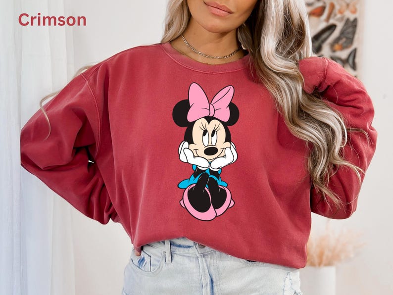 May include: Crimson-colored sweatshirt featuring a Minnie Mouse graphic. The graphic shows Minnie Mouse with a pink bow, blue accents, and black ears. The word "Crimson" is in the upper left corner. The sweatshirt has long sleeves.