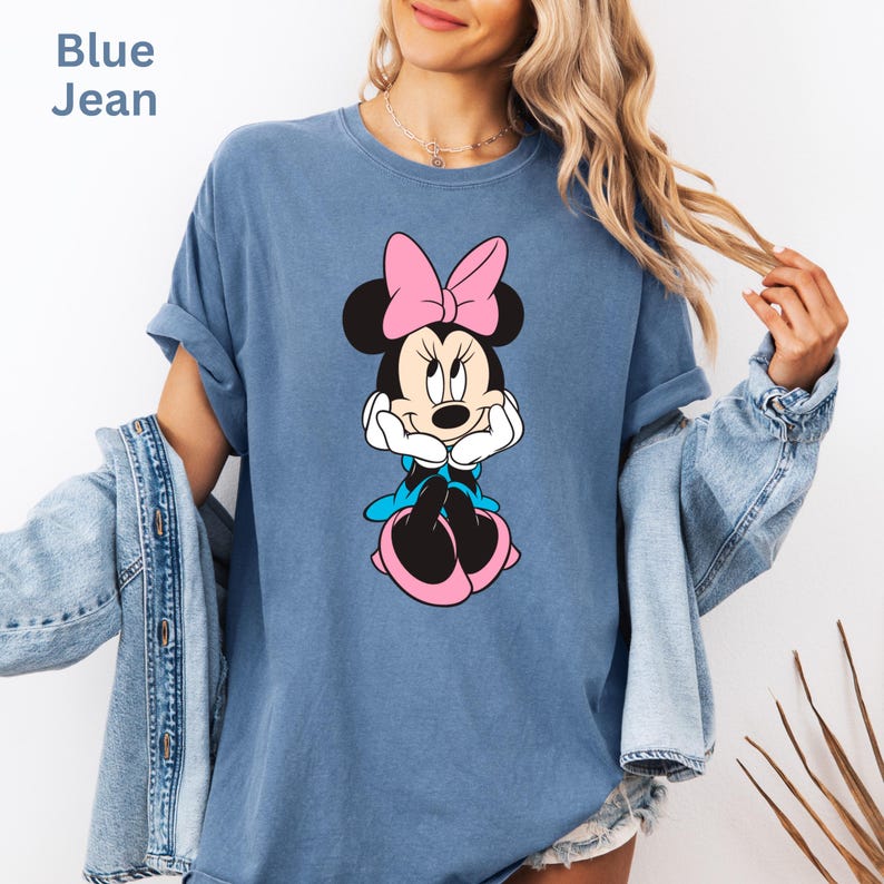 May include: Blue jean colored t-shirt featuring a Minnie Mouse graphic. The graphic shows Minnie Mouse with a pink bow, black ears, and a blue dress. The text "Blue Jean" is in the upper left corner.