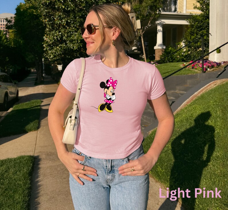 May include: Light pink t-shirt featuring a Minnie Mouse graphic. The cartoon character is depicted with a pink bow, dress, and black and yellow shoes. The shirt is paired with blue jeans. The text "Light Pink" is visible at the bottom of the image.