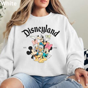 May include: White sweatshirt featuring the word "Disneyland" in a black, stylized font, with the text "EST. 1955". The sweatshirt showcases a colorful graphic of Disney characters, including Mickey and Minnie Mouse, Donald Duck, and Goofy.