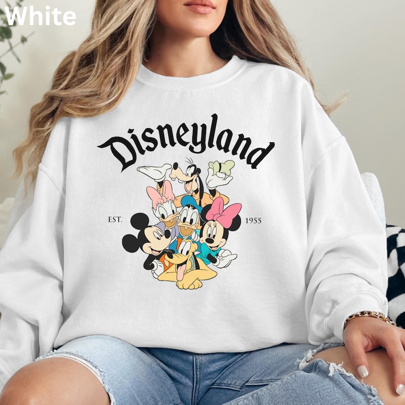 May include: White sweatshirt featuring the word "Disneyland" in a black, stylized font, with the text "EST. 1955". The sweatshirt showcases a colorful graphic of Disney characters, including Mickey and Minnie Mouse, Donald Duck, and Goofy.