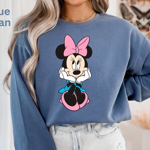 May include: Blue jean colored sweatshirt featuring a Minnie Mouse graphic. The graphic shows Minnie Mouse with a pink bow, black ears, and a white face. The sweatshirt has long sleeves and a crew neck. The text "Blue Jean" is in the upper left corner.