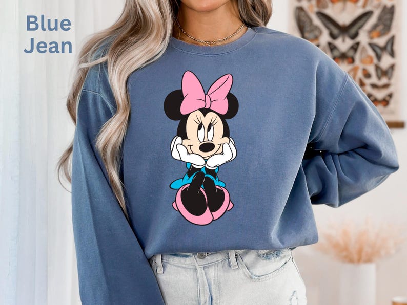 May include: Blue jean colored sweatshirt featuring a Minnie Mouse graphic. The graphic shows Minnie Mouse with a pink bow, black ears, and a white face. The sweatshirt has long sleeves and a crew neck. The text "Blue Jean" is in the upper left corner.