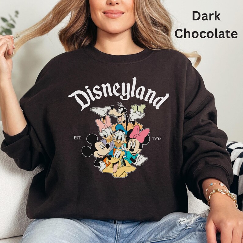 May include: Dark chocolate-colored sweatshirt featuring the word "Disneyland" in white script above a graphic of Disney characters. The sweatshirt includes the text "EST. 1955".