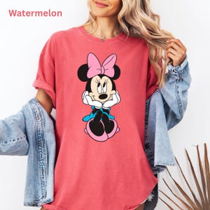 May include: A watermelon-colored t-shirt featuring a Minnie Mouse graphic. The cartoon character is depicted with a pink bow, black ears, and a blue dress. The word "Watermelon" is printed in the upper left corner.