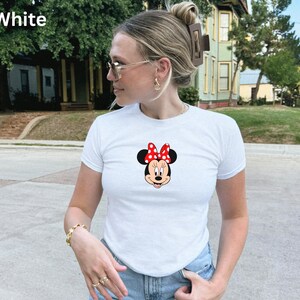 May include: White t-shirt featuring a Minnie Mouse graphic. The graphic shows Minnie's face with a red bow with white polka dots. The t-shirt is short-sleeved and appears to be made of a soft material. The word "White" is in the top left corner.