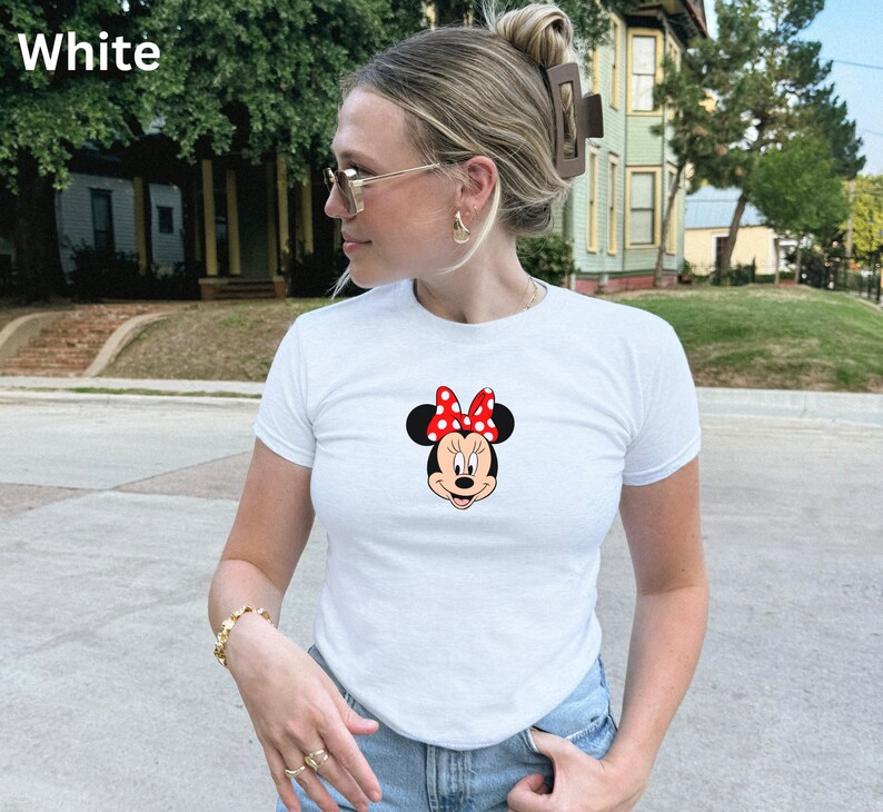 May include: White t-shirt featuring a Minnie Mouse graphic. The graphic shows Minnie's face with a red bow with white polka dots. The t-shirt is short-sleeved and appears to be made of a soft material. The word "White" is in the top left corner.