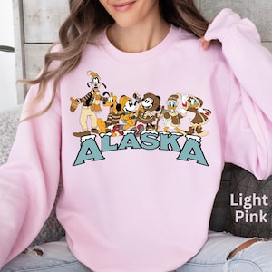 Peut inclure: Sweat-shirt rose clair avec un motif de personnages de dessins anim&eacute;s en v&ecirc;tements d'hiver. Le mot "ALASKA" est imprim&eacute; en grandes lettres bleu clair sur le devant. Le sweat-shirt est de couleur rose clair unie.