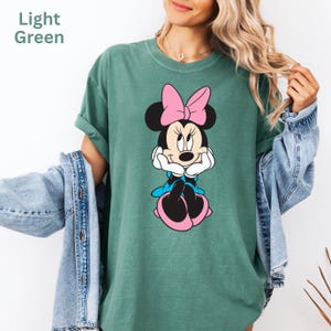 May include: Light green t-shirt featuring a Minnie Mouse graphic. The cartoon character is depicted with a pink bow, blue dress, and pink shoes. The text "Light Green" is in the upper left corner. The shirt is a casual style.