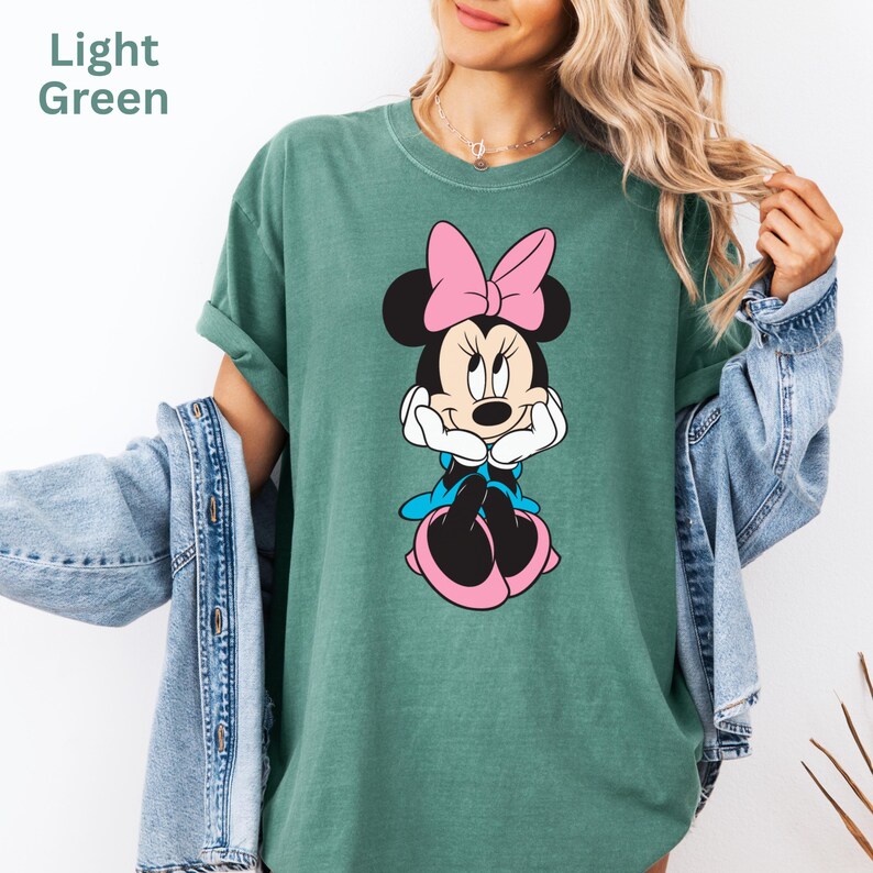 May include: Light green t-shirt featuring a Minnie Mouse graphic. The cartoon character is depicted with a pink bow, blue dress, and pink shoes. The text "Light Green" is in the upper left corner. The shirt is a casual style.